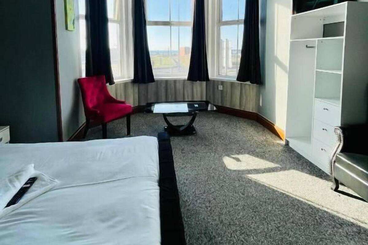 A hotel room with a bed, a red chair, a small table, a white wardrobe, and a large window with curtains.