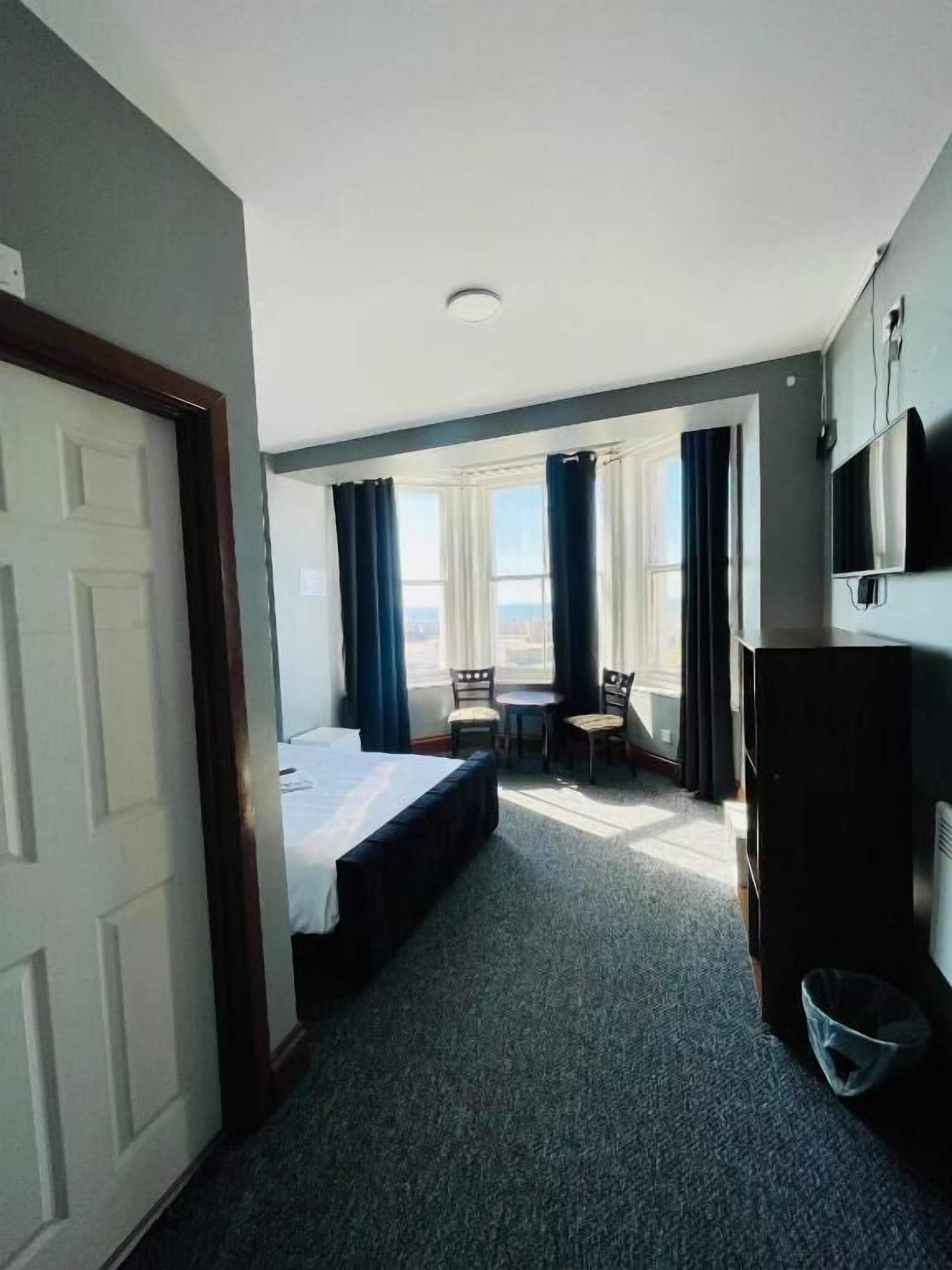 A hotel room with a bed, a desk with chairs, a TV, and a window with curtains.