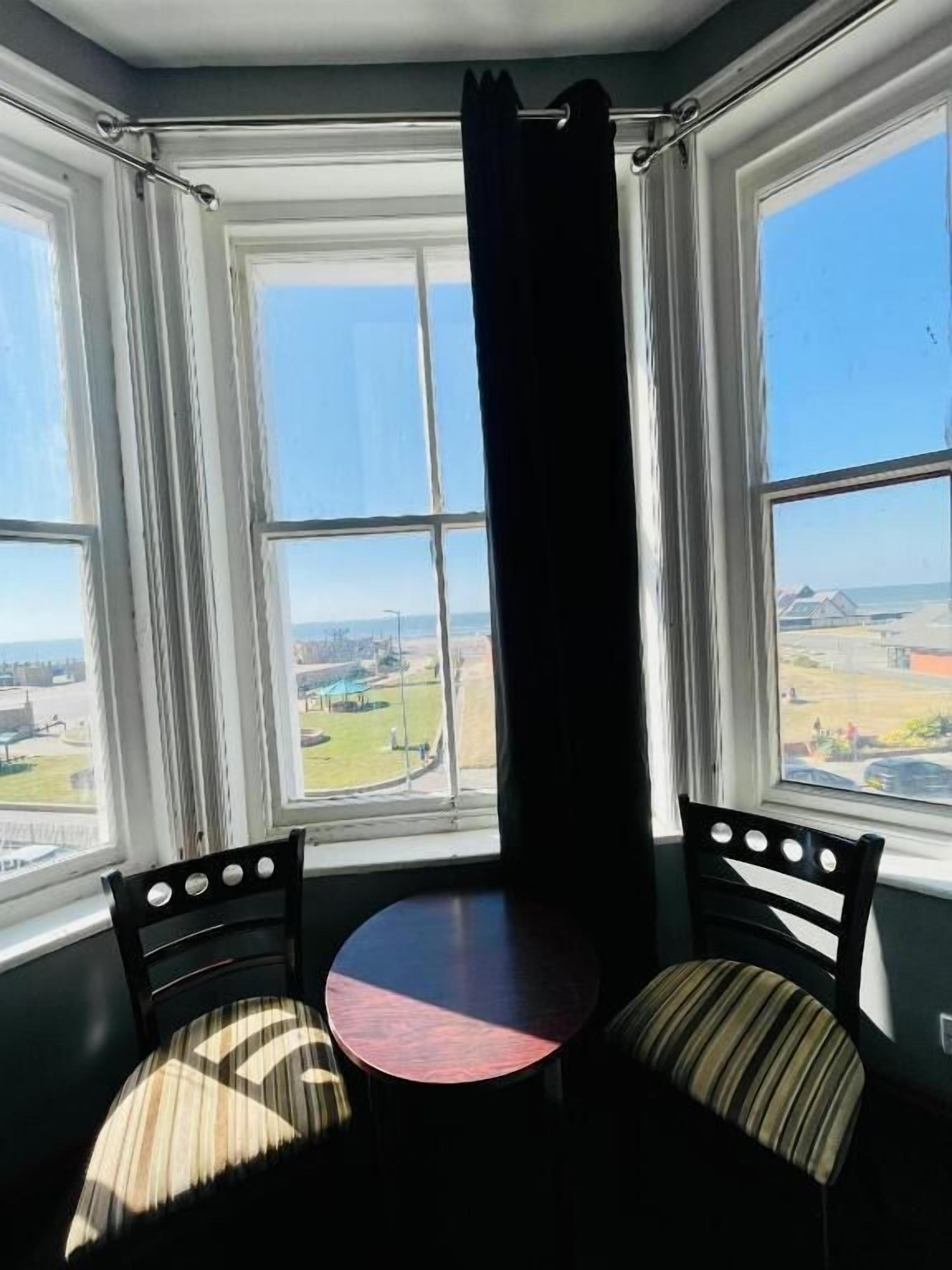 Ein Eckzimmer mit Erkerfenster, zwei Sessel, einem kleinen Tisch und Blick auf eine Landschaft.