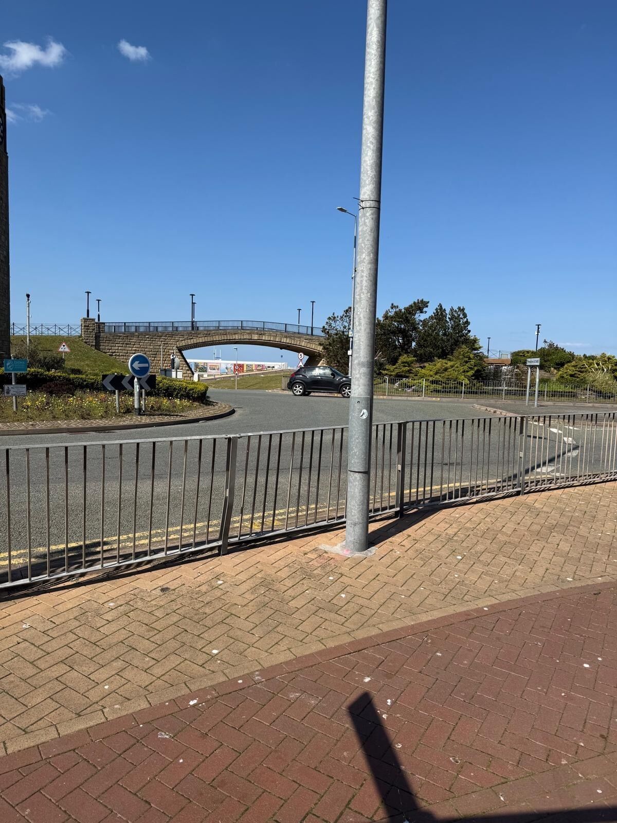 Uma rotatória com cerca de metal, caminho para pedestres e uma ponte ao fundo.