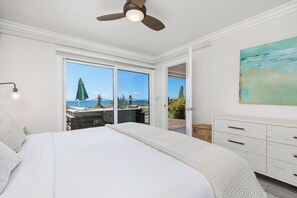 Ein Schlafzimmer mit einem großen Bett, einem Deckenventilator, Meerblick durch Schiebetüren und einer weißen Kommode.