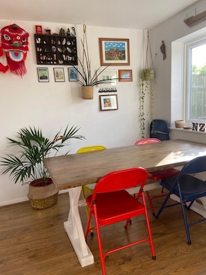 A dining area with a wooden table, red and blue chairs, a red lion head decoration, a wall-mounted plant, and a framed picture. - Two minutes walk from beach. Located on quiet pathway. Dogs welcome. (Camber)