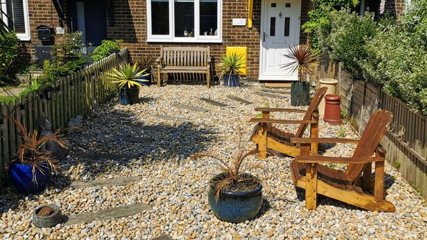 Une maison à deux étages, construite en briques, avec un toit recouvert de tuiles, une porte d’entrée et un petit jardin agrémenté d’un banc, de plantes en pot et d’une clôture en bois.