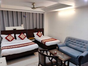 A hotel room with two beds, a blue armchair, a small table, and a ceiling fan. - Hotel Ratandeep @ Paharganj (New Delhi)