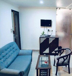 A living room with a sofa, a glass-top coffee table, a chair, a TV mounted on the wall, and a cabinet with a plant on it. - Hotel Ratandeep @ Paharganj (New Delhi)