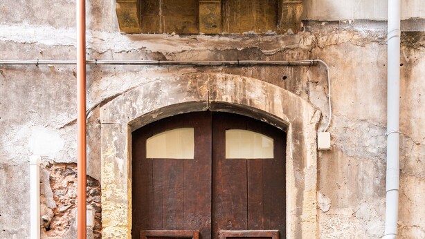 Una puerta de madera rústica con un pequeño ventanal, un muro de piedra y una tubería de metal.