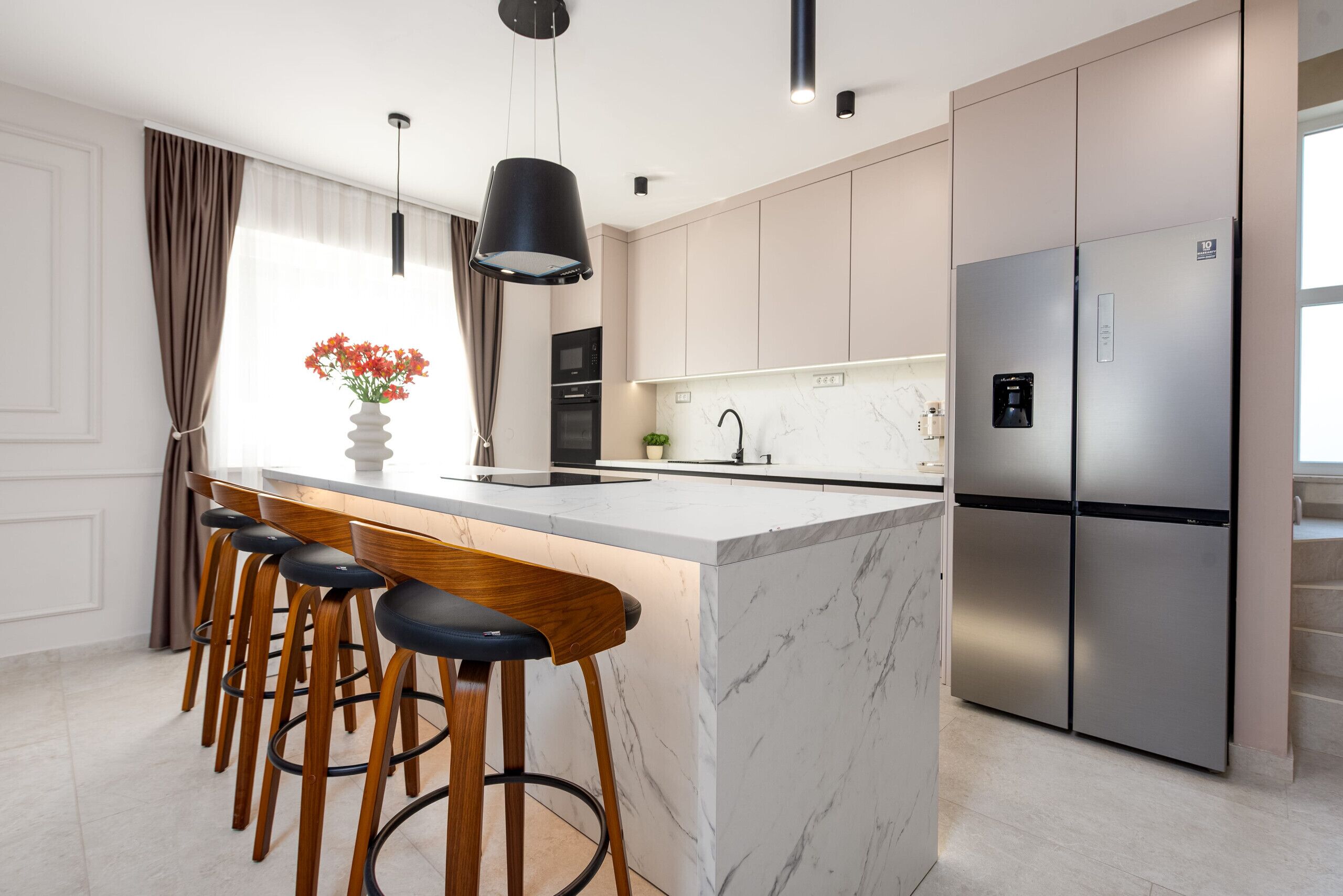 Une cuisine moderne dotée d’un îlot en marbre, de tabourets de bar, d’appareils électroménagers en acier inoxydable et de luminaires suspendus.