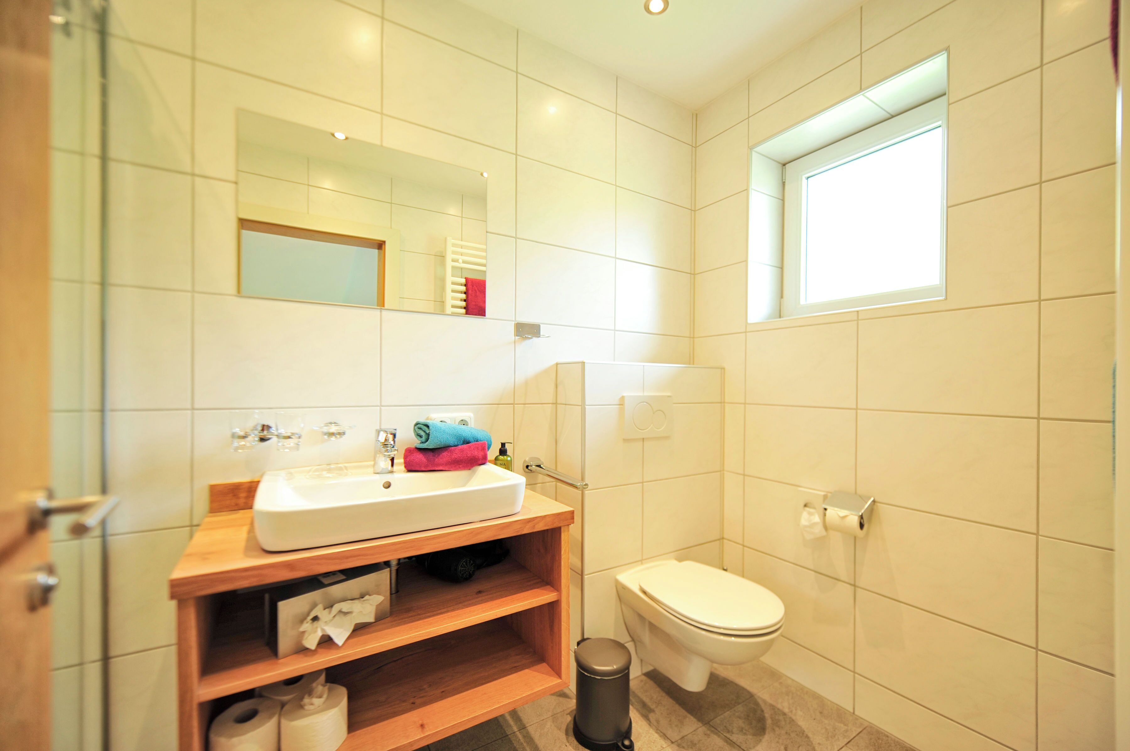 A bathroom with a white sink, mirror, and toilet, wooden shelves, and a window.