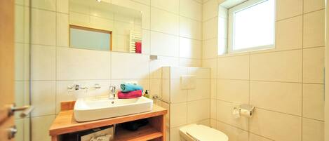 A bathroom with a white sink, mirror, and toilet, wooden shelves, and a window.