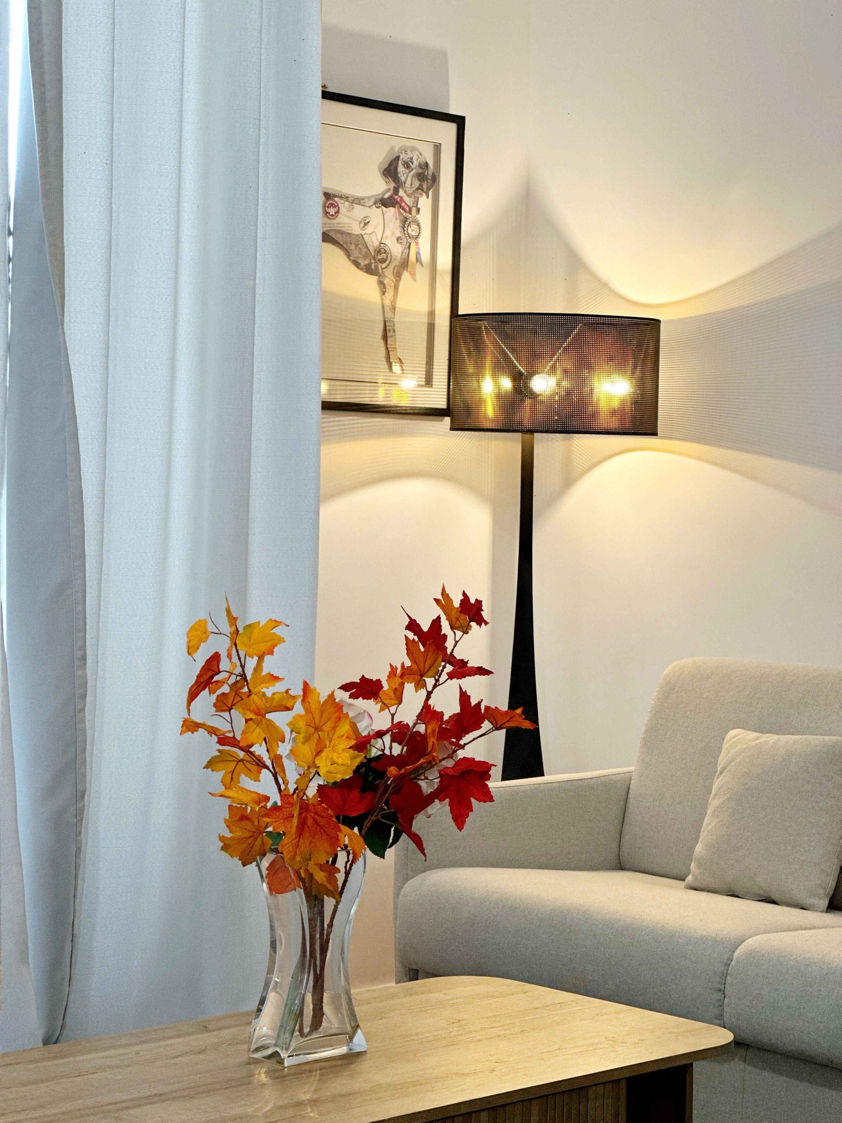 A living room with a sofa, a table with a vase of autumn leaves, a framed picture on the wall, and a floor lamp.