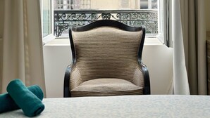 Una habitación con una silla cerca de una ventana, una cama con una colcha estampada y una toalla enrollada sobre la cama.