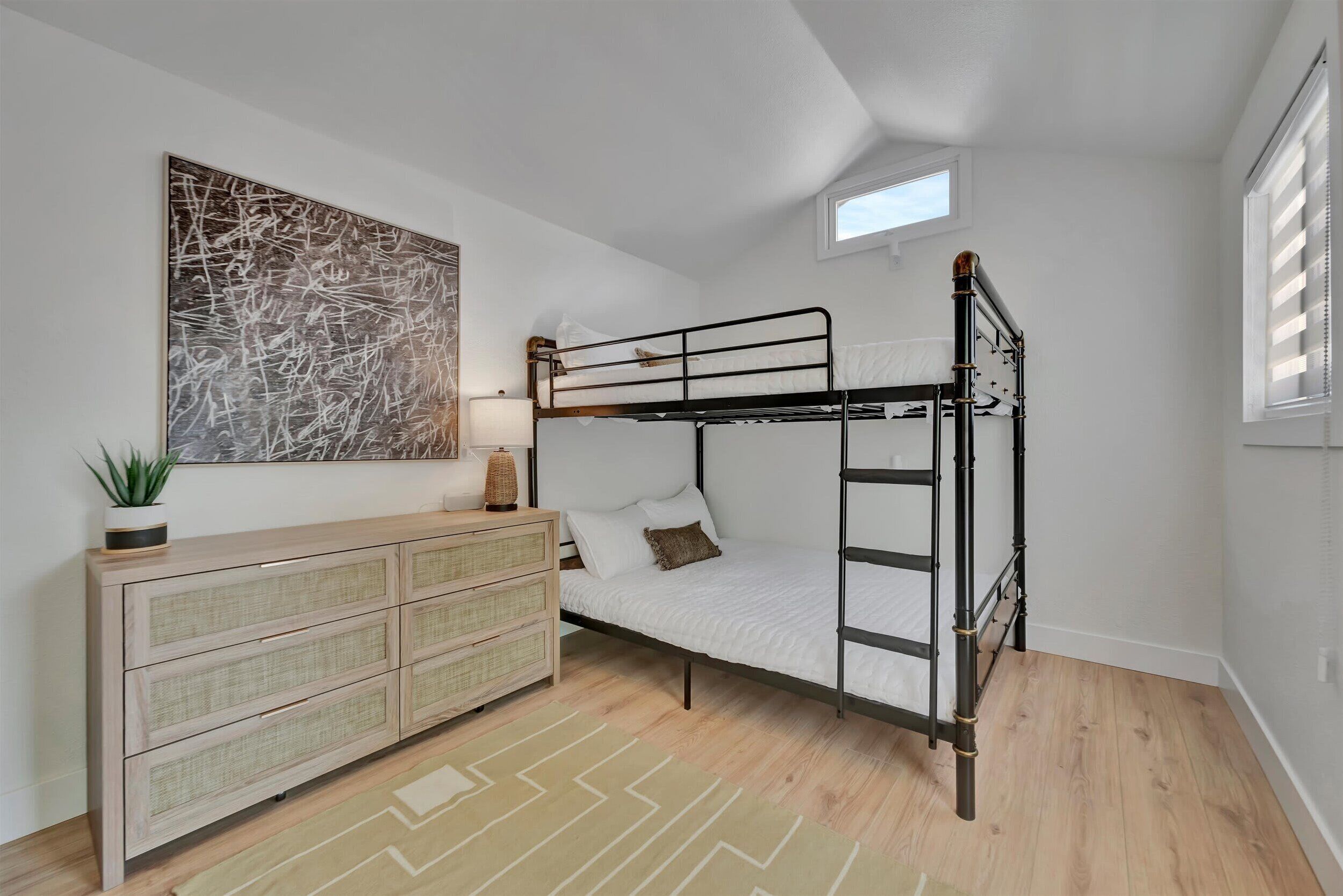 Une chambre avec un lit superposé, une commode en rotin, une œuvre d’art murale et une fenêtre avec des stores.