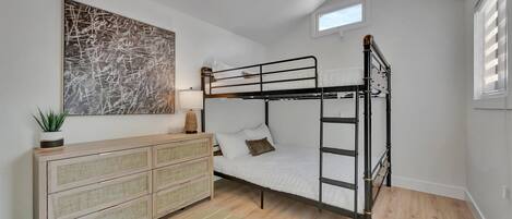 Une chambre avec un lit superposé, une commode en rotin, une œuvre d’art murale et une fenêtre avec des stores.