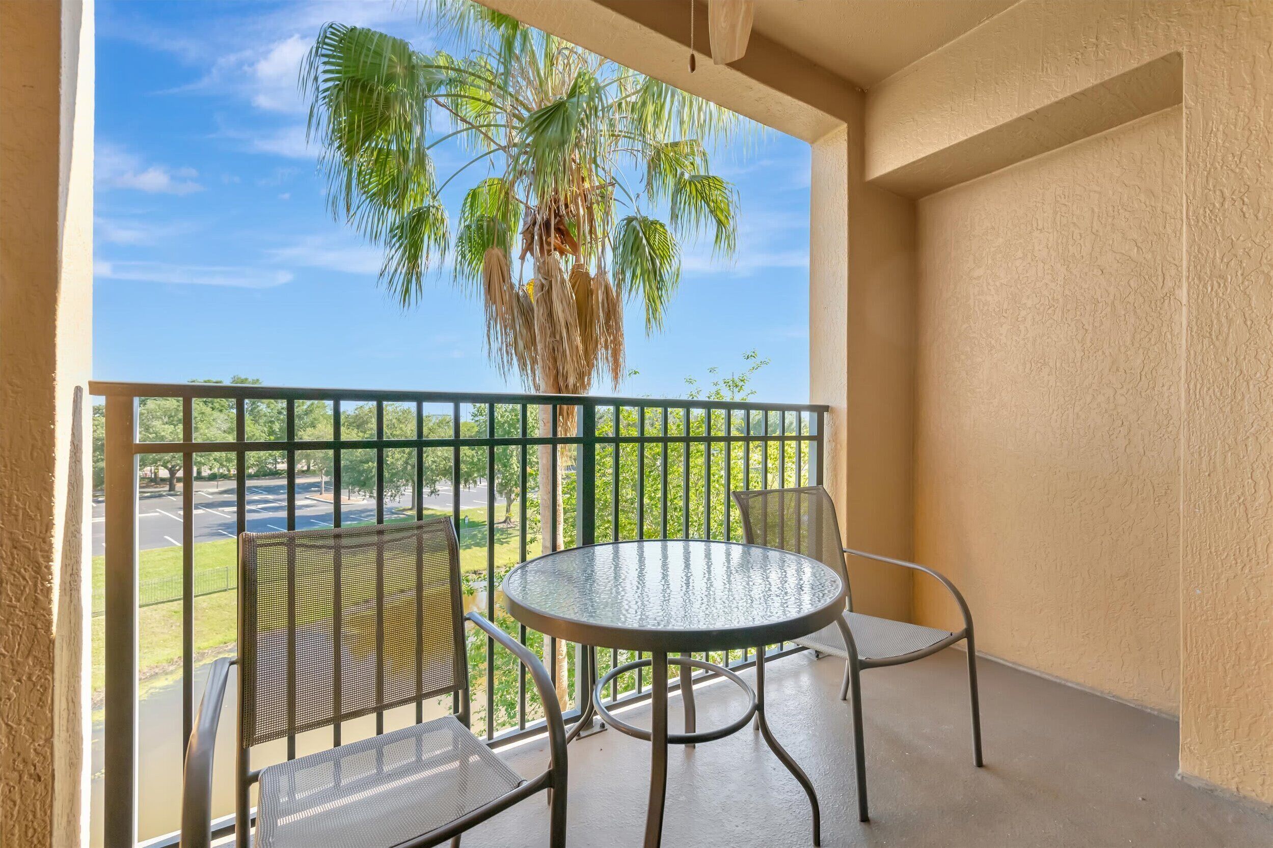 A balcony with a round table and two chairs, a palm tree, and a view of a parking lot and greenery.