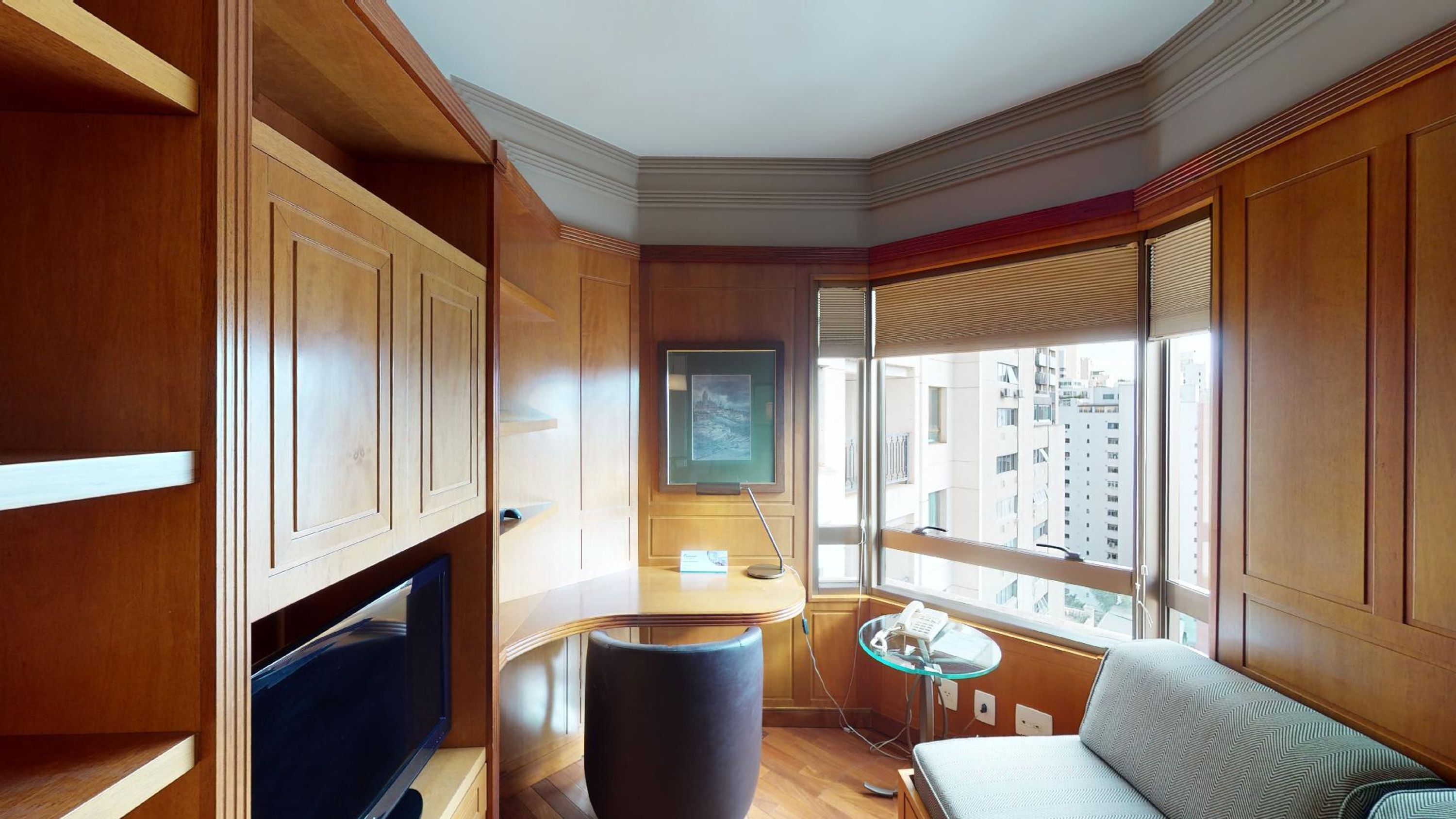 A corner room with wooden paneling, a television, a glass table with a telephone, and a window with a city view.