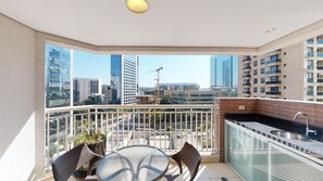 Ein Balkon mit Glastür, einem kleinen Tisch mit Stühlen, einem Blumentopf und Blick auf Stadtgebäude.