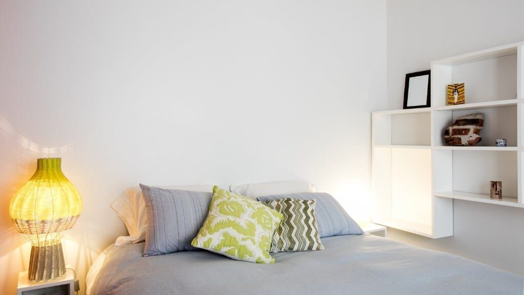 A bedroom with a bed, pillows, a bedside lamp, and a white shelving unit against the wall.