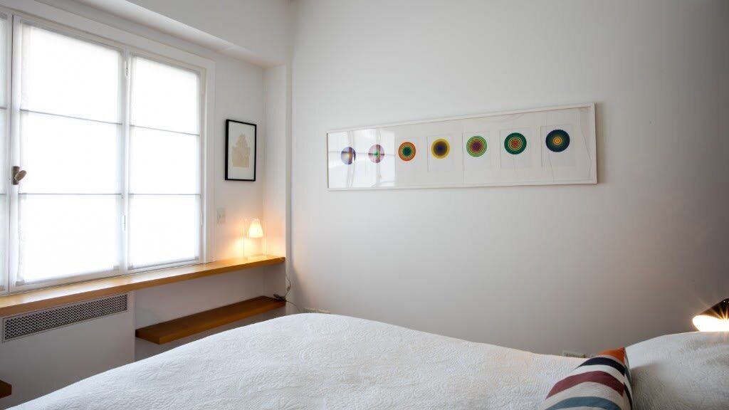 A bedroom with a large window, a bedside table, a bed, and a wall-mounted artwork.