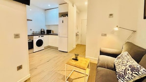 A compact living space with a kitchenette, a sofa with a patterned pillow, and a small wooden table. - Republica 2BR apt Near Madrid s Financial Center (Madrid)