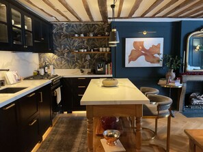A modern kitchen with dark cabinetry, a central island, and a dining area with a table and chairs. - Elegance Marais A Distinctive High end Marais one Bedroom (Paris)