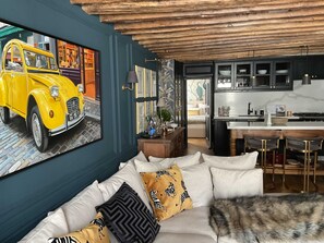 A modern living room with a sofa, pillows, a painting of a vintage car, and a kitchen area with cabinets and a dining table. - Elegance Marais A Distinctive High end Marais one Bedroom (Paris)