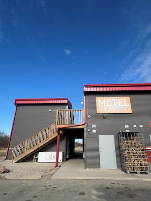 Un motel con un letrero que indica que vende cerveza, una escalera que sube a un segundo nivel y una pila de leña afuera.
