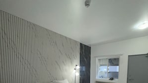 A bedroom with a bed, bedside table, wall-mounted air conditioning unit, window, and a door.