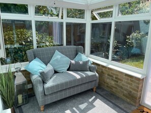 Ein Wintergarten mit einem grauen, karierten Sofa, zwei Kissen und einem Glastisch mit einer Kerze.