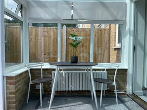 Eine überdachte Terrasse mit Tisch und zwei Stühlen, einem Blumentopf und Blick auf einen Holenzaun.