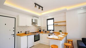 Een compacte keuken met witte kastjes, een houten aanrecht, een gasfornuis, een vaatwasser en een raam. Er staan krukken bij het aanrecht en een zwarte bank in de hoek.