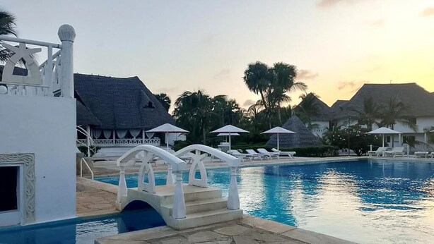 Piscina com uma ponte branca, cadeiras de descanso e palmeiras ao fundo.