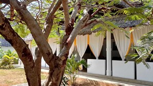 Uma trilha arborizada leva a um edifício com telhado de palha e cortinas brancas.
