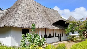 Um edifício com telhado de palha, fachada branca, uma pequena varanda e um jardim com plantas tropicais.