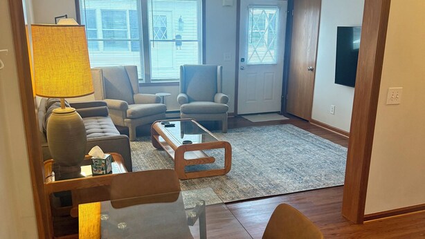 A living room with a glass dining table, two armchairs, a sofa, a lamp, and a television. - Find comfort in this cozy, recently updated apartment. Featuring two bedrooms, a full kitchen with oak cabinets, and a generous living space. It's designed for easy living with a ranch-style, step-up entry. Located in a peaceful neighborhood. (Marion)