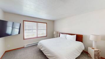 A hotel room with a bed, a flat-screen TV, a window with blinds, and a nightstand with a lamp.