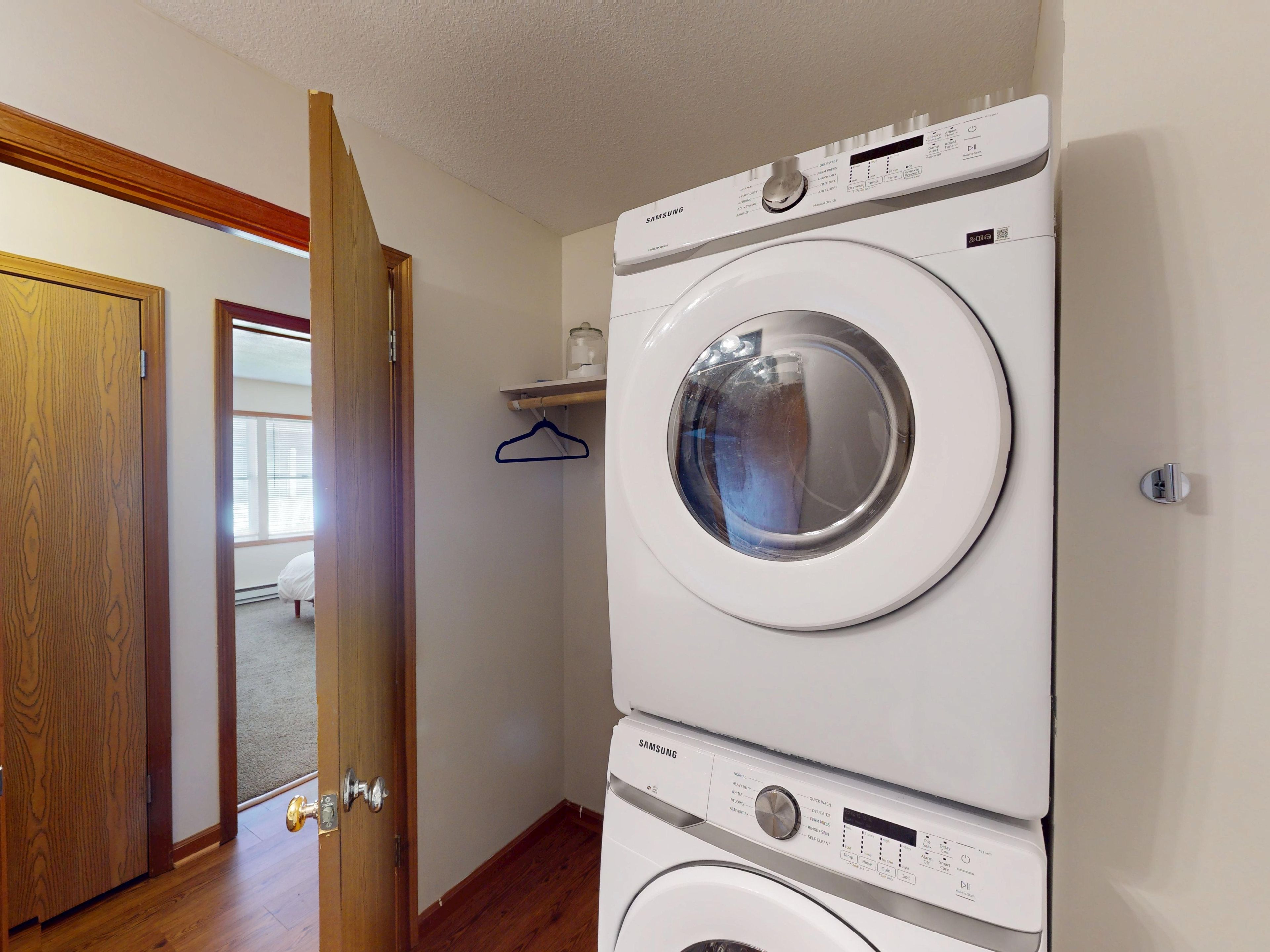 A laundry room with stacked washers and dryers, wooden flooring, a door leading to another room, and a small shelf with a hanging item.