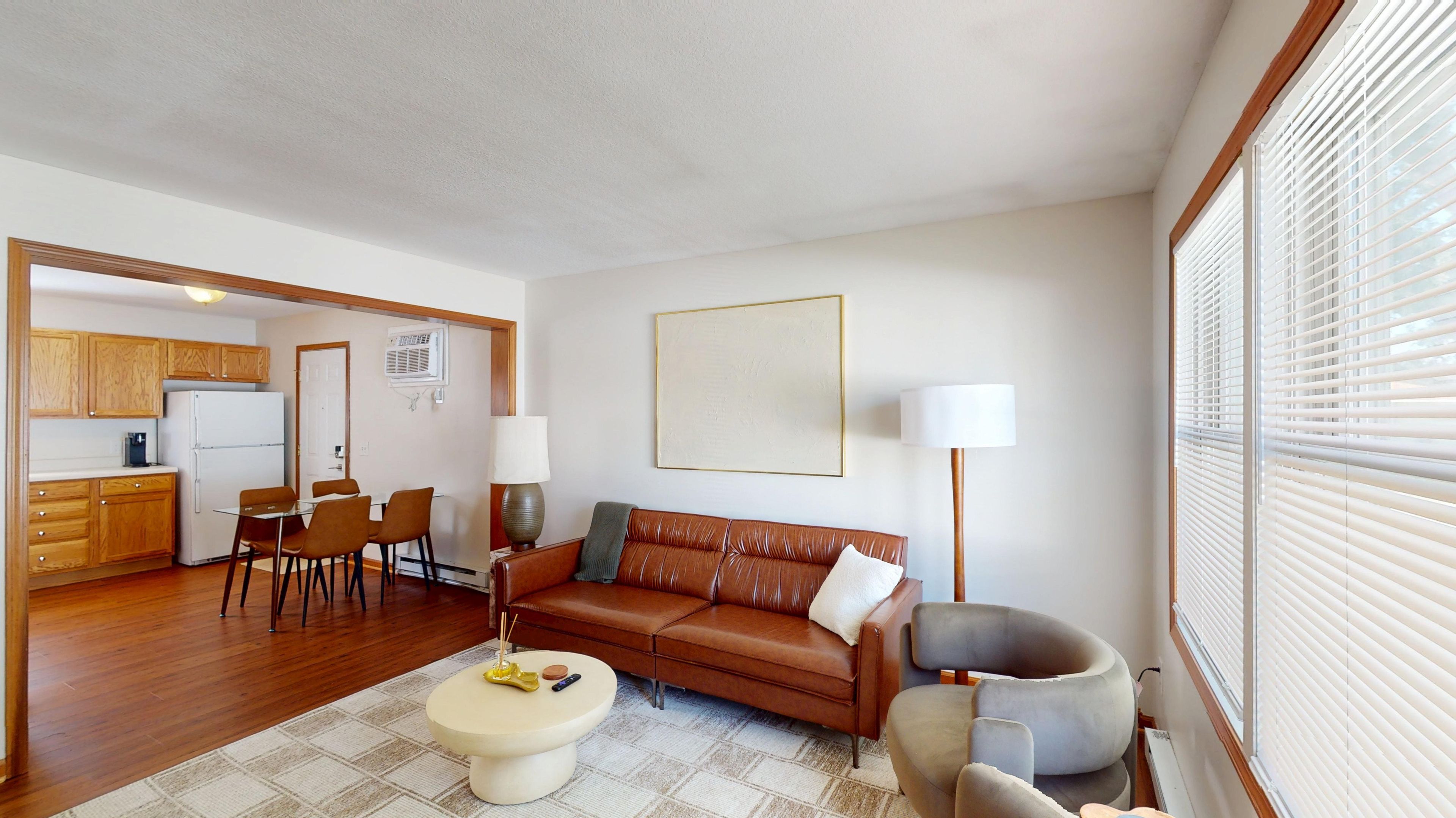 A living room with a brown leather sofa, a coffee table, and a dining area with wooden cabinets and a white refrigerator.