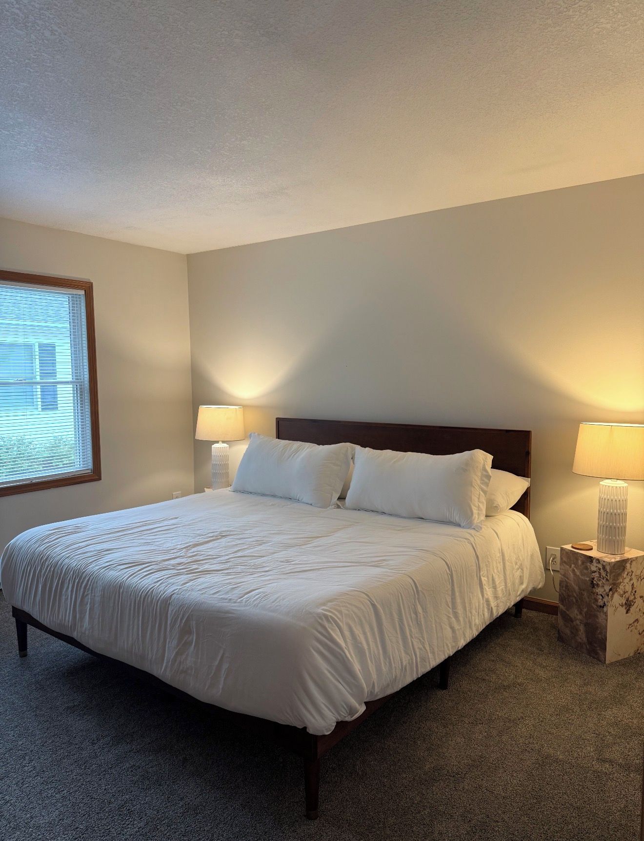 A neatly made bed with white bedding, two bedside lamps, and a window with blinds.