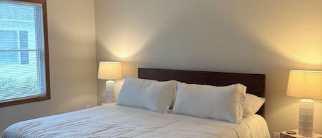 A neatly made bed with white bedding, two bedside lamps, and a window with blinds.
