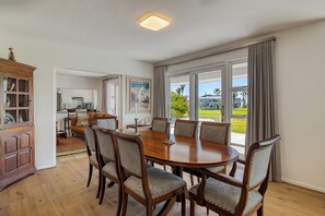 Un coin repas avec une table et des chaises en bois, une cuisine à l’arrière-plan et de grandes fenêtres avec des rideaux.