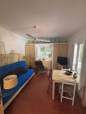 Una habitación con un sofá azul, una mesa de comedor de madera, una silla estampada, un ventilador de techo y un televisor.