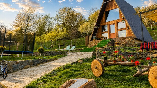 A wooden A-frame house with a stone foundation, a stone pathway, and a hammock.