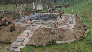 A stone pathway leading to a circular seating area with a fire pit, surrounded by a rustic garden with potted plants and a small pond.