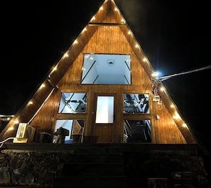 A triangular wooden building with a central entrance, multiple windows, and string lights. - Căsuța de pe Vale (Sighetu Marmației)