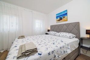 Ein ordentlich bezogenes Bett mit einem gemusterten Überwurf, ein Strandbild an der Wand und ein Nachttisch mit einer Lampe.