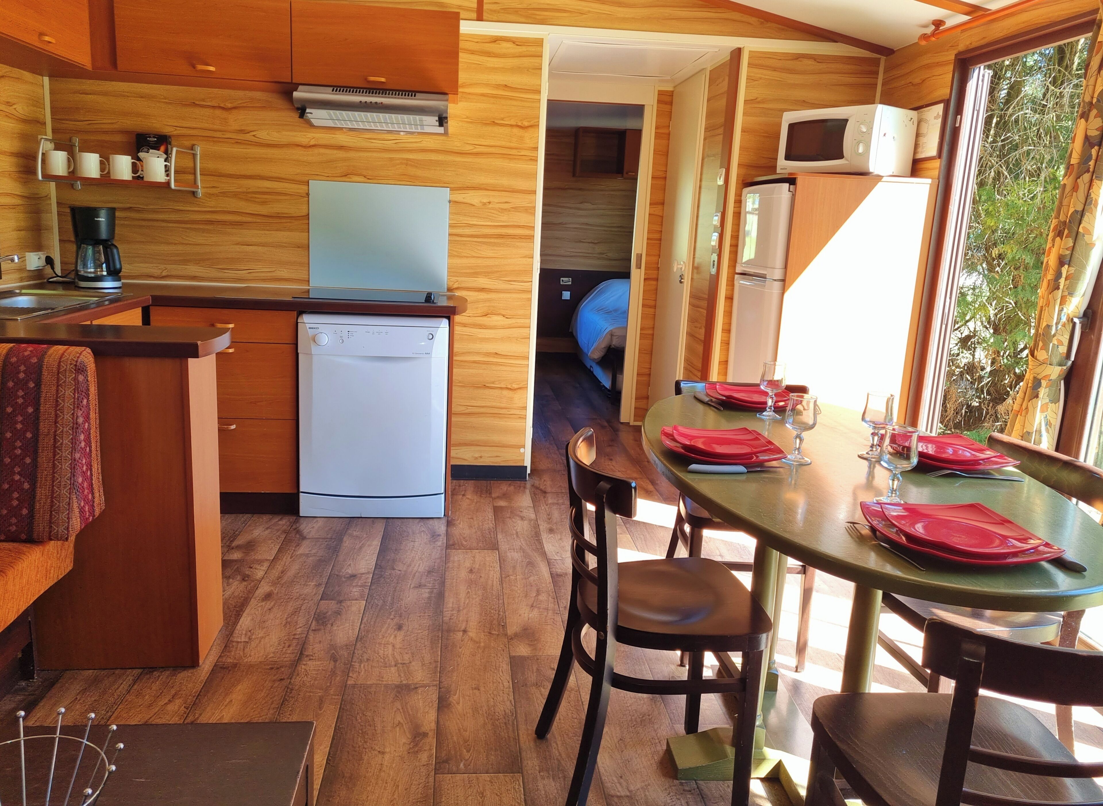 A compact kitchen with a dishwasher, a microwave, and a coffee maker. There is a dining area with a table set for two, and a view of the outdoors through a window.