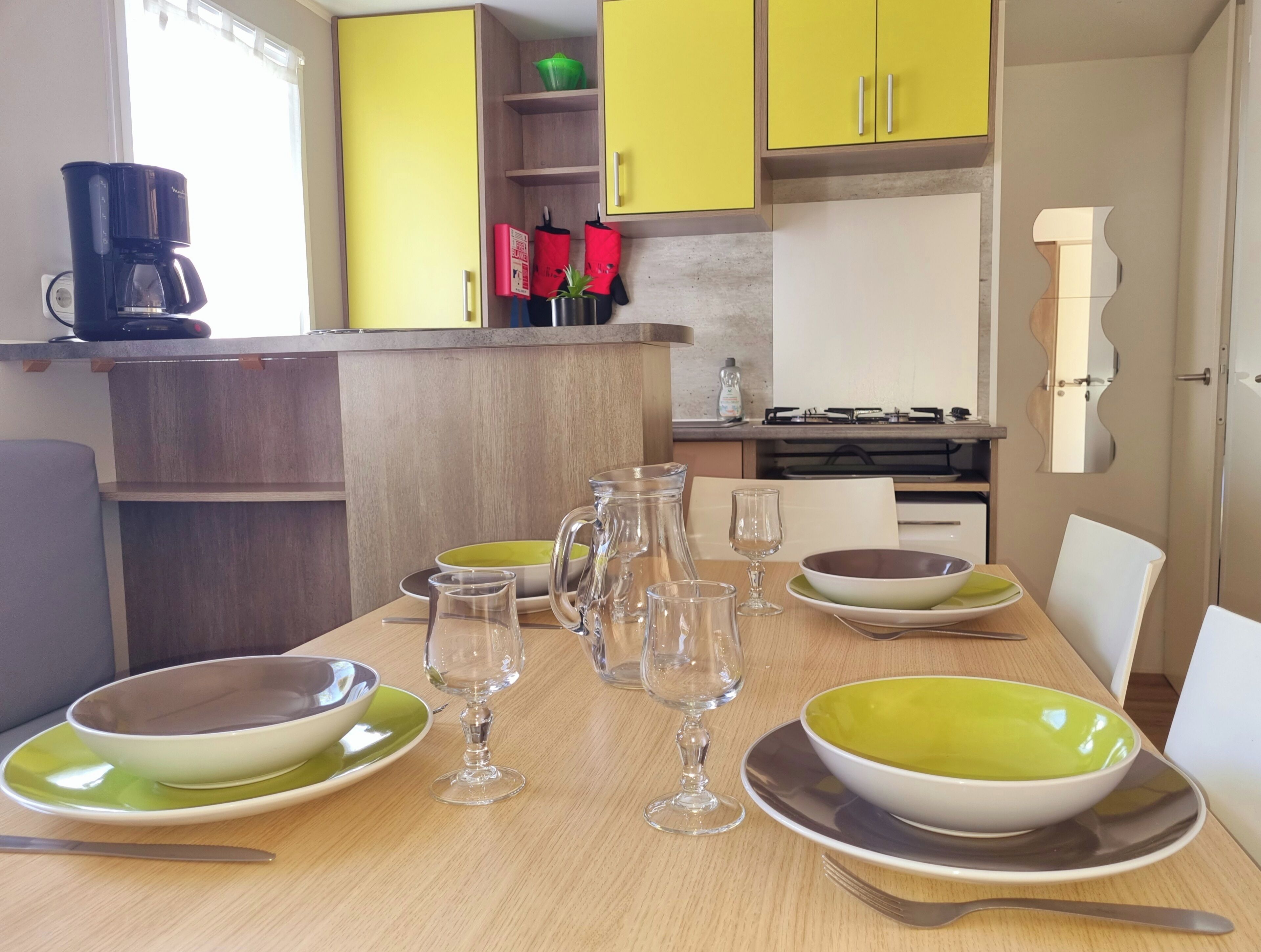 Une cuisine moderne avec des meubles de couleur jaune, une machine à café et une table à manger agrémentée de bols verts et blancs, de verres à vin et de couverts.