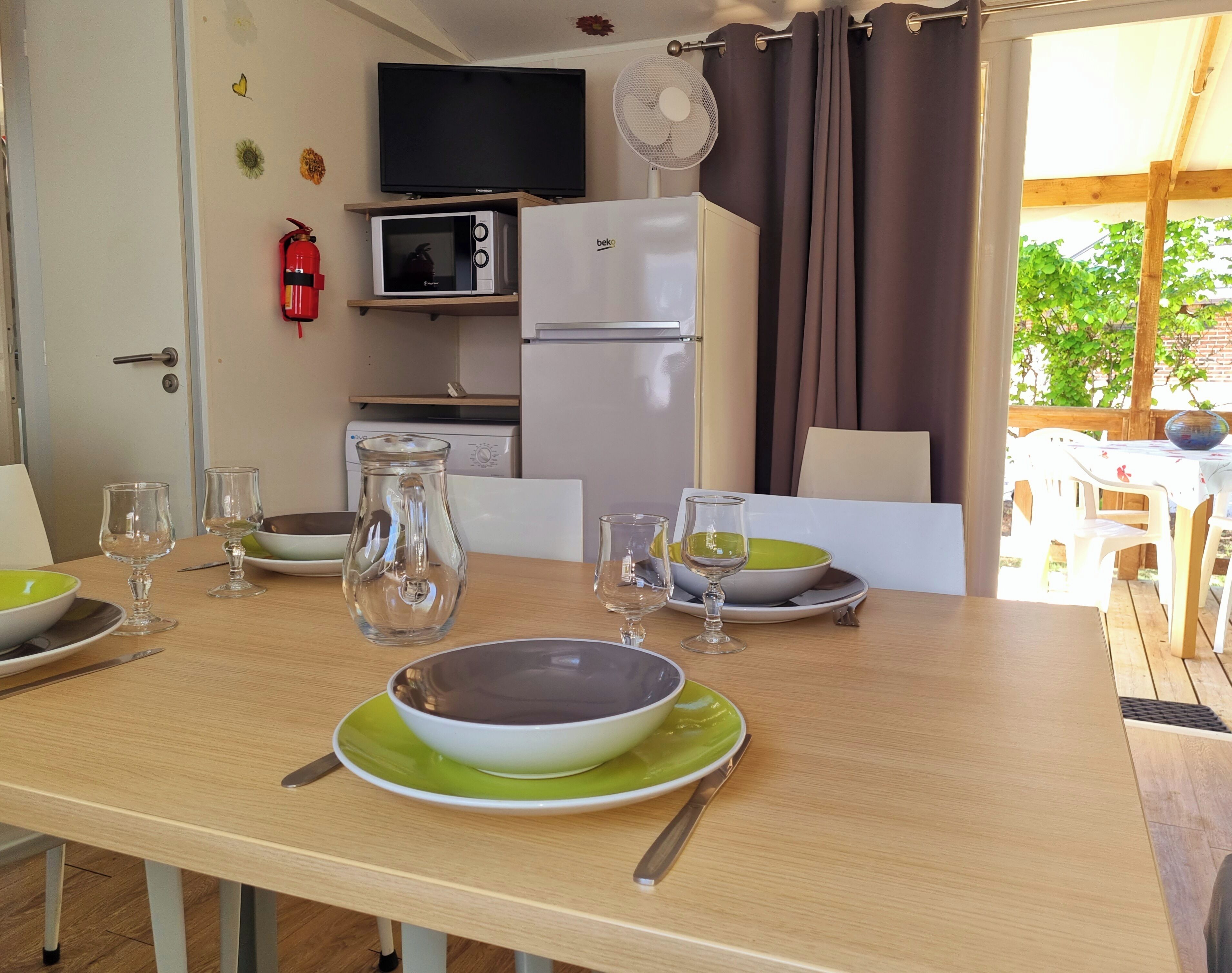 Una mesa de comedor lista con platos, tazones y vasos, un microondas, un refrigerador, un televisor y un extintor en una habitación con una puerta y una ventana.