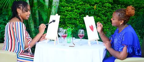 Dos personas pintando sobre caballetes a una mesa con copas de vino y una botella verde.