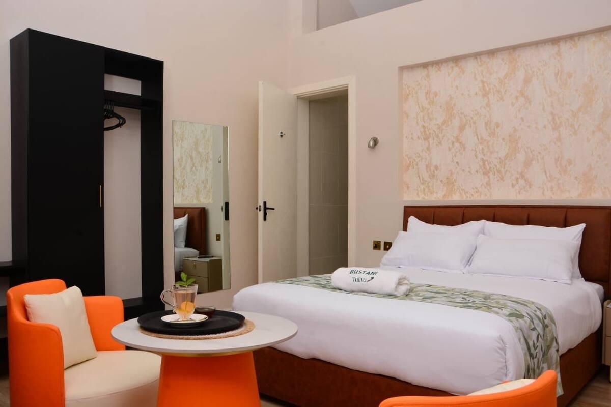 A hotel room with a bed, a small table with a drink, an orange chair, and a wardrobe.